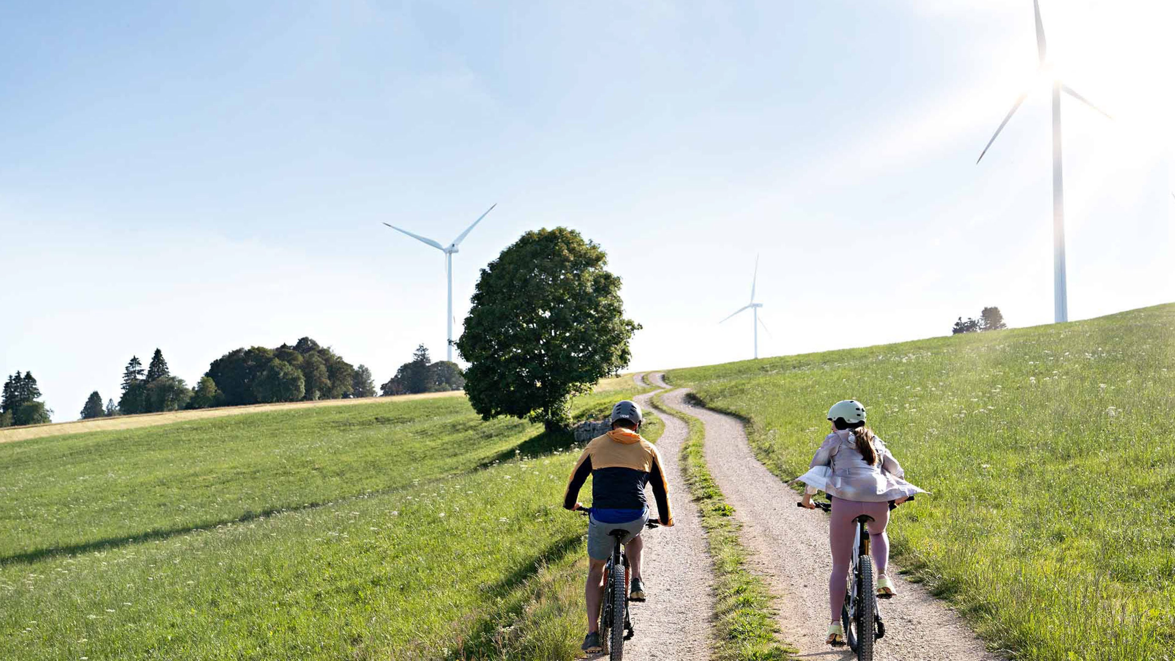 Two people riding bicycles along a dirt road, with windwheels in the background.