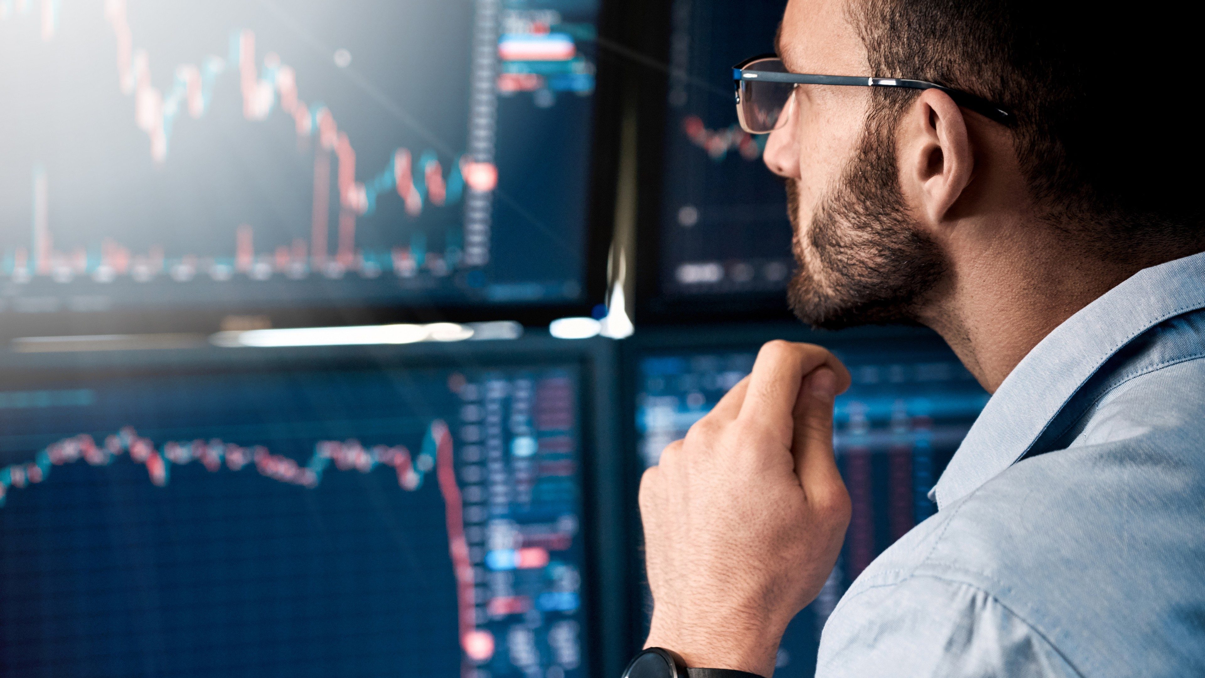 Bearded man trader wearing eyeglasses sitting at desk at office monitoring stock market looking at monitors analyzing candle bar price flow touching chin concerned trading concept close-up; Shutterstock ID 1803009610; purchase_order: -; job: -; client: -; other: -