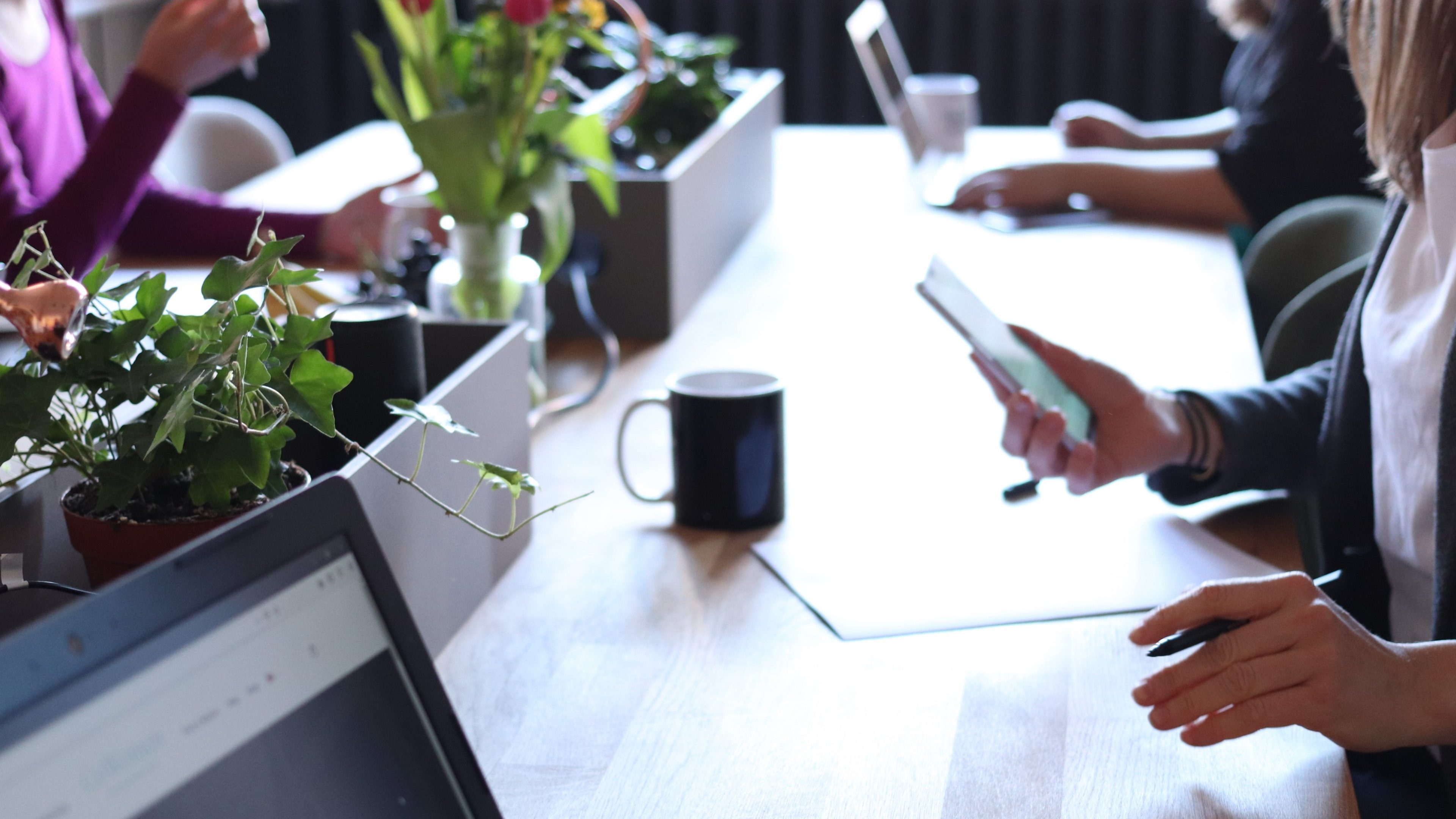 Espace de travail collaboratif avec des plantes vertes, des tasses de café et des personnes utilisant des appareils numériques autour d'une grande table.