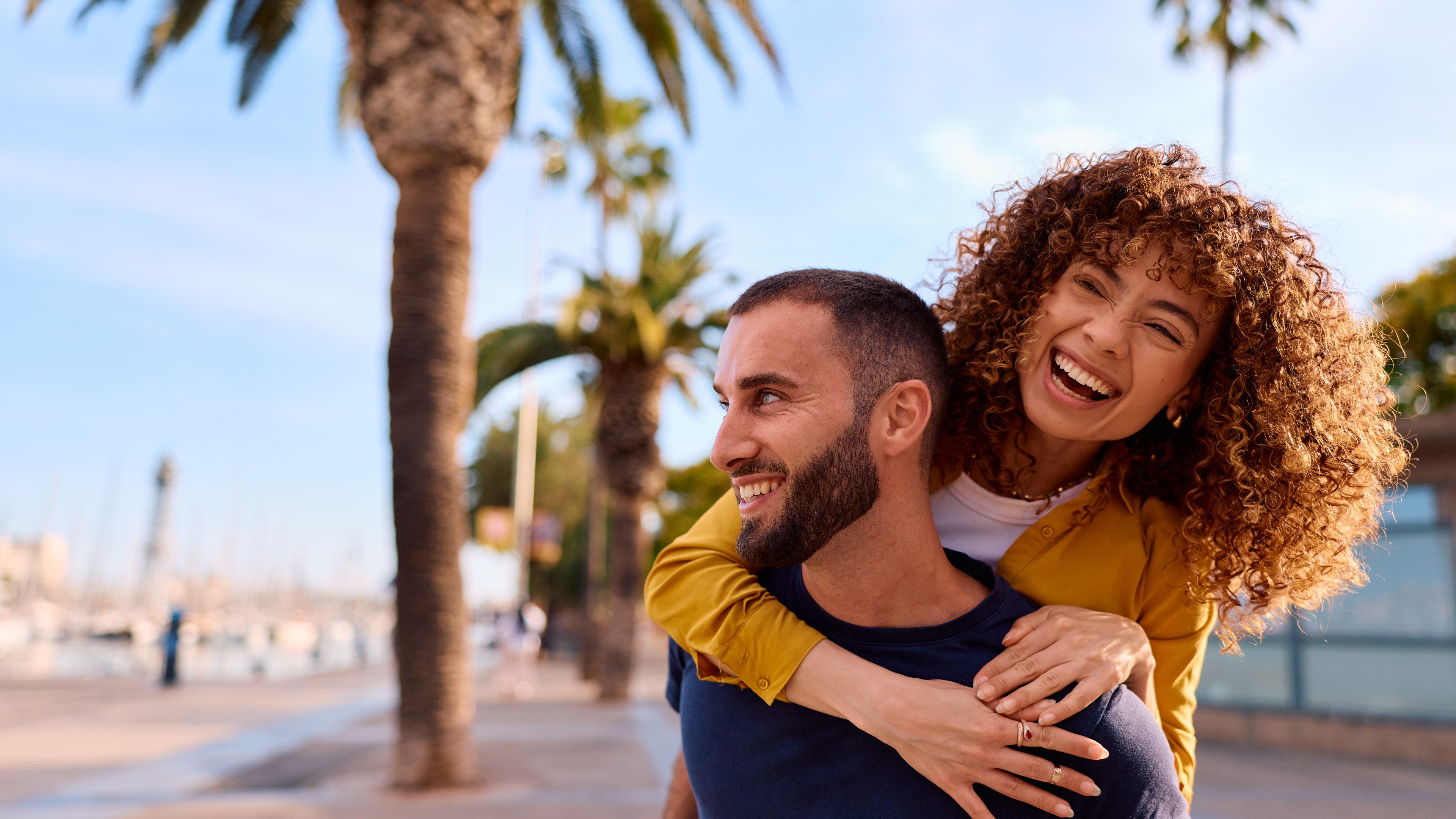 Happy young adult woman laughing while receiving a piggyback ride from a man on a sunny day with palm trees