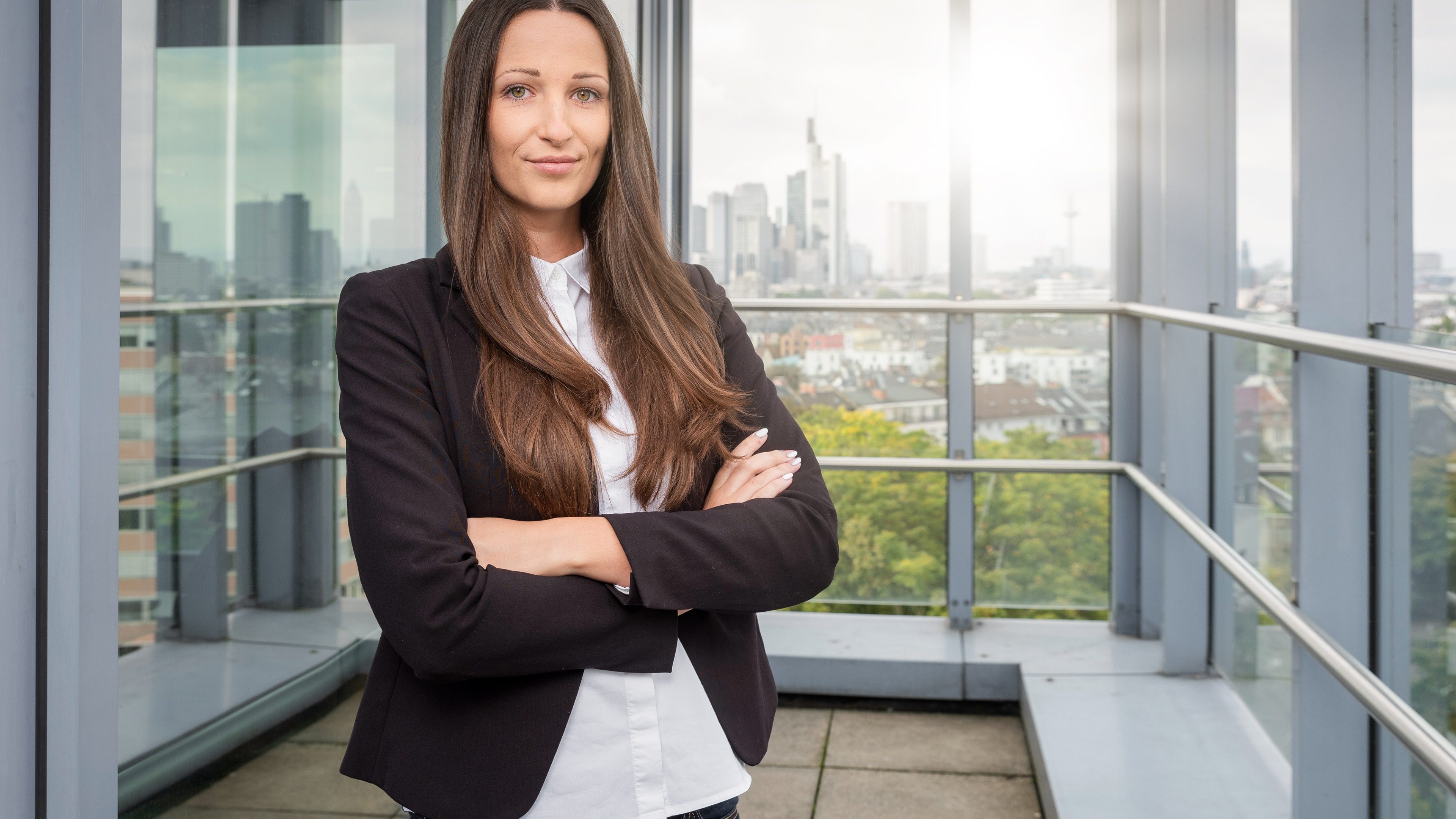 Portrait d'une jeune professionnelle souriante, bras croisés, posant sur une terrasse de bureau avec une vue panoramique sur un quartier d'affaires moderne, symbolisant l'ambition et la réussite.