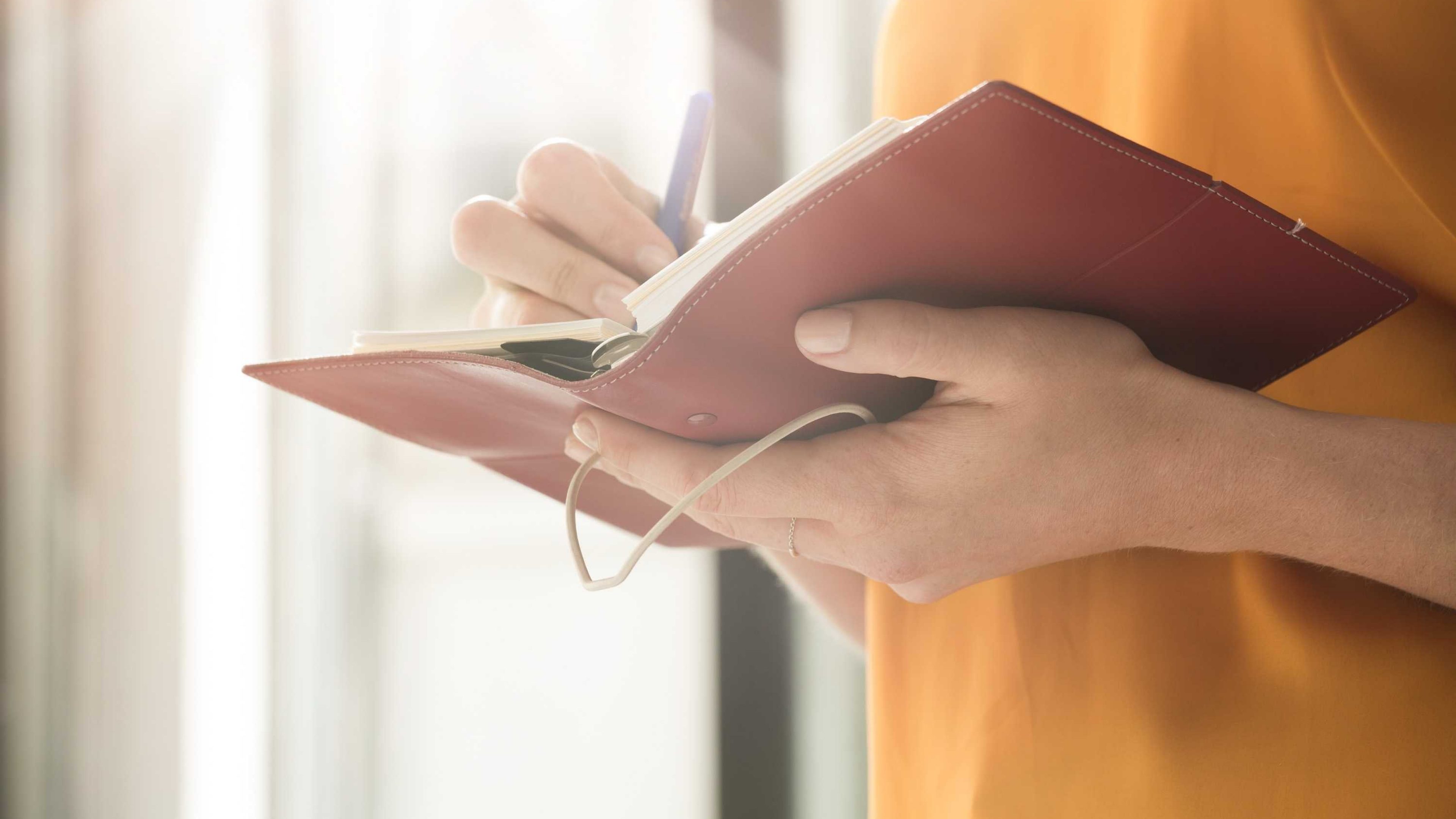 Gros plan sur les mains d'une personne prenant des notes dans un carnet de bord en cuir rouge, symbolisant l'analyse et l'étude des marchés.