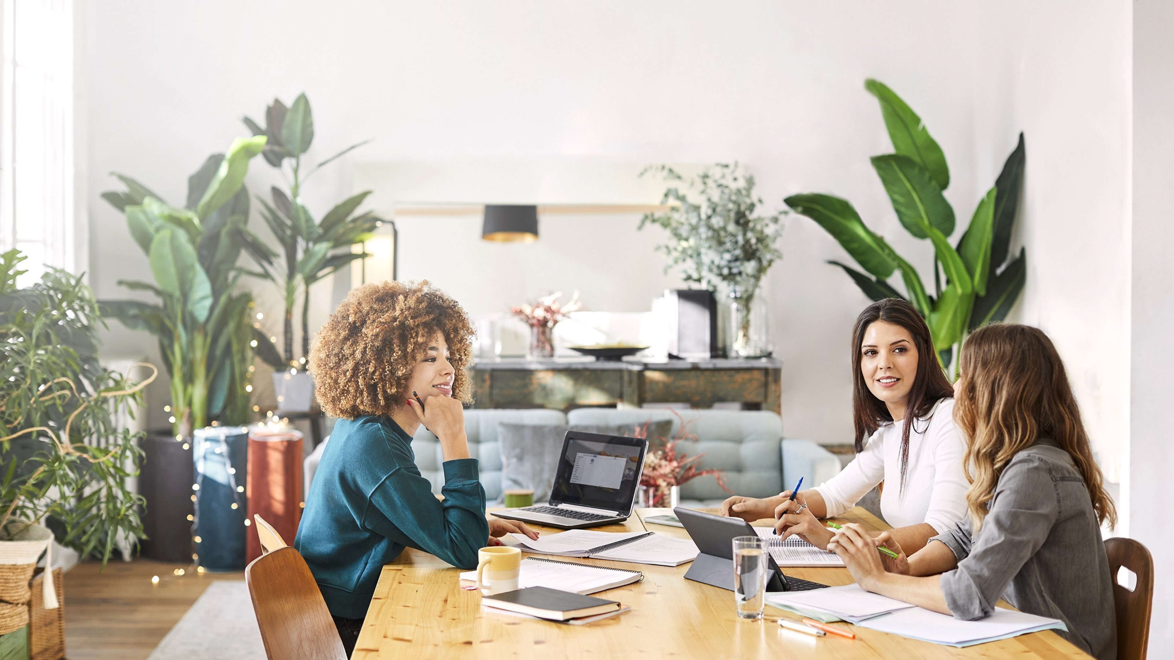 Trois professionnelles en réunion de travail collaborative autour d'une table boisée, utilisant une tablette et des documents papier dans un environnement de bureau chaleureux et moderne.