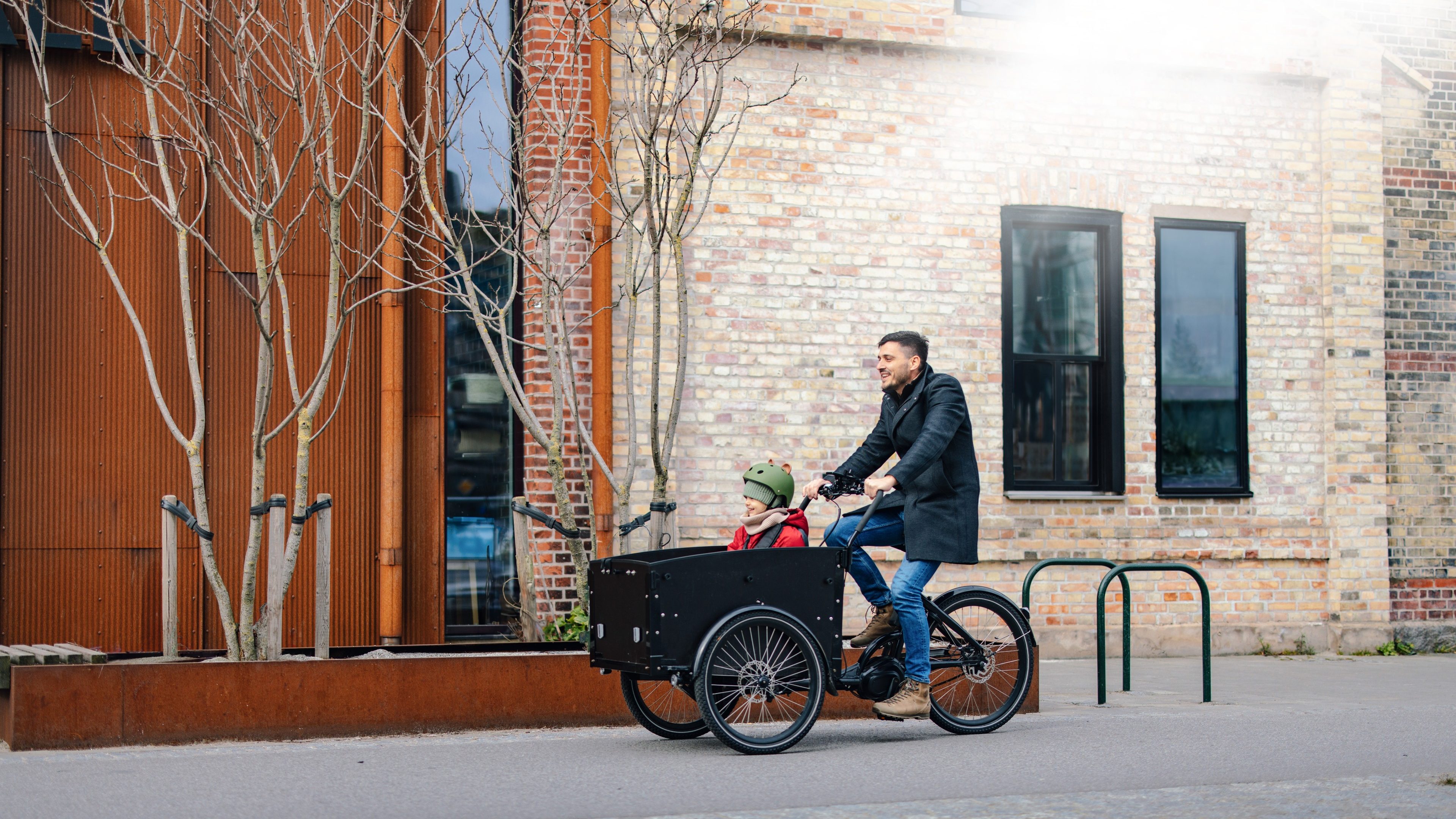 Un père transportant son enfant dans un vélo-cargo devant un immeuble de bureaux en briques, illustrant l'engagement du fonds SLF Dynapierre ELTIF dans l'immobilier durable et la vie urbaine de demain.