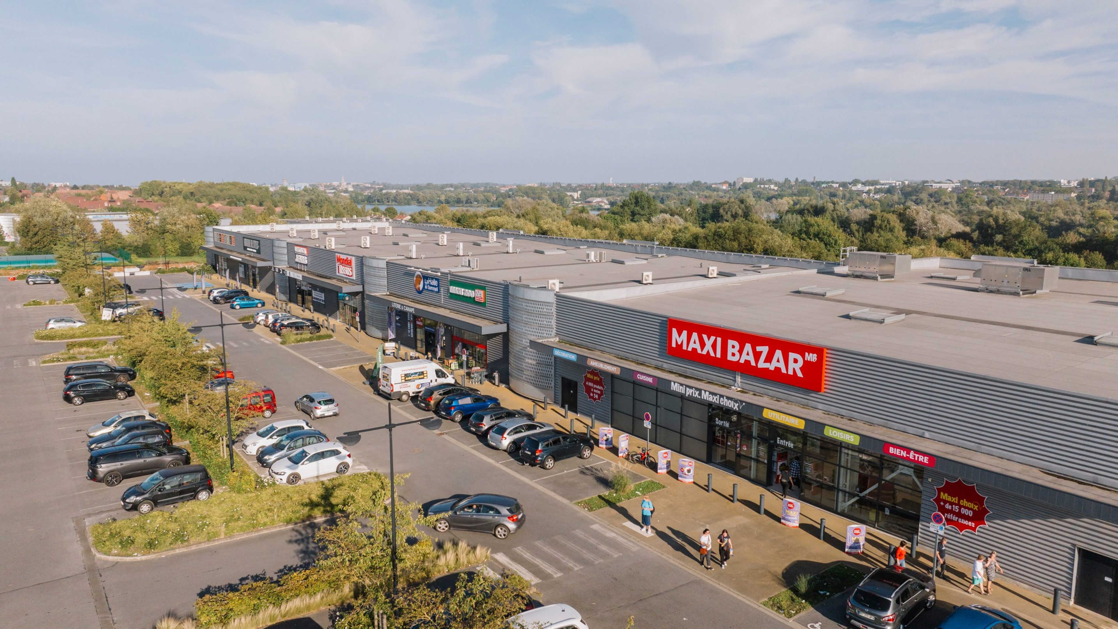 Vue aérienne d’un parc commercial avec plusieurs enseignes, un parking en façade et un magasin Maxibazar au premier plan
