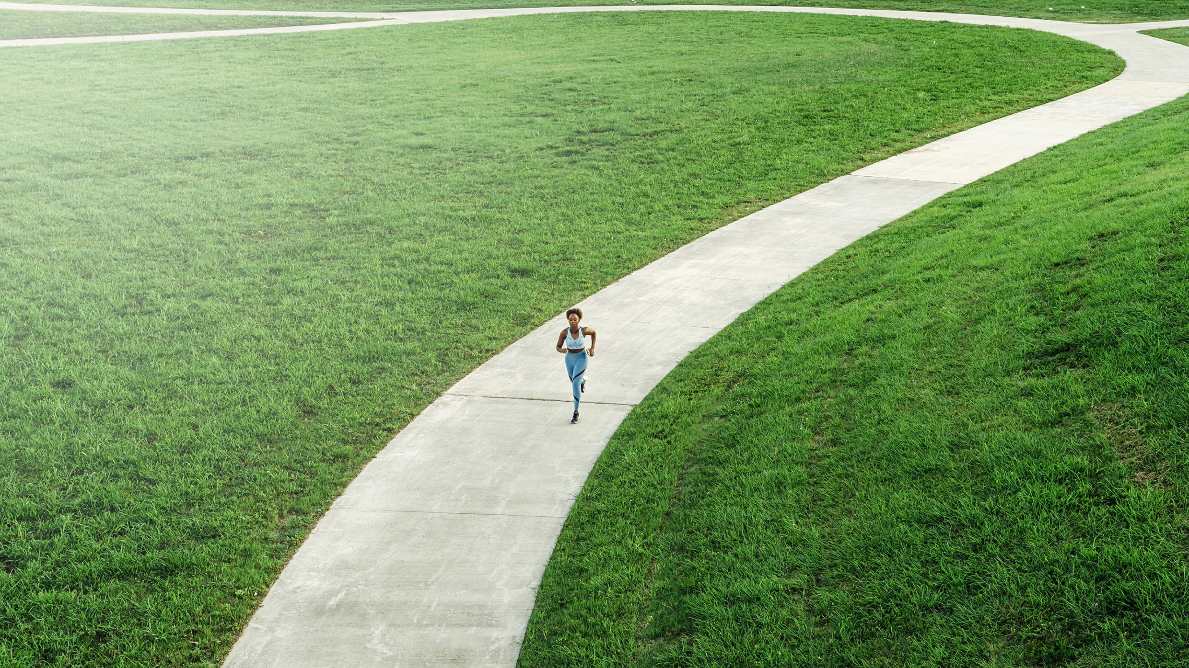 Vue aérienne d'une femme en tenue de sport courant sur un chemin sinueux traversant un vaste parc verdoyant.