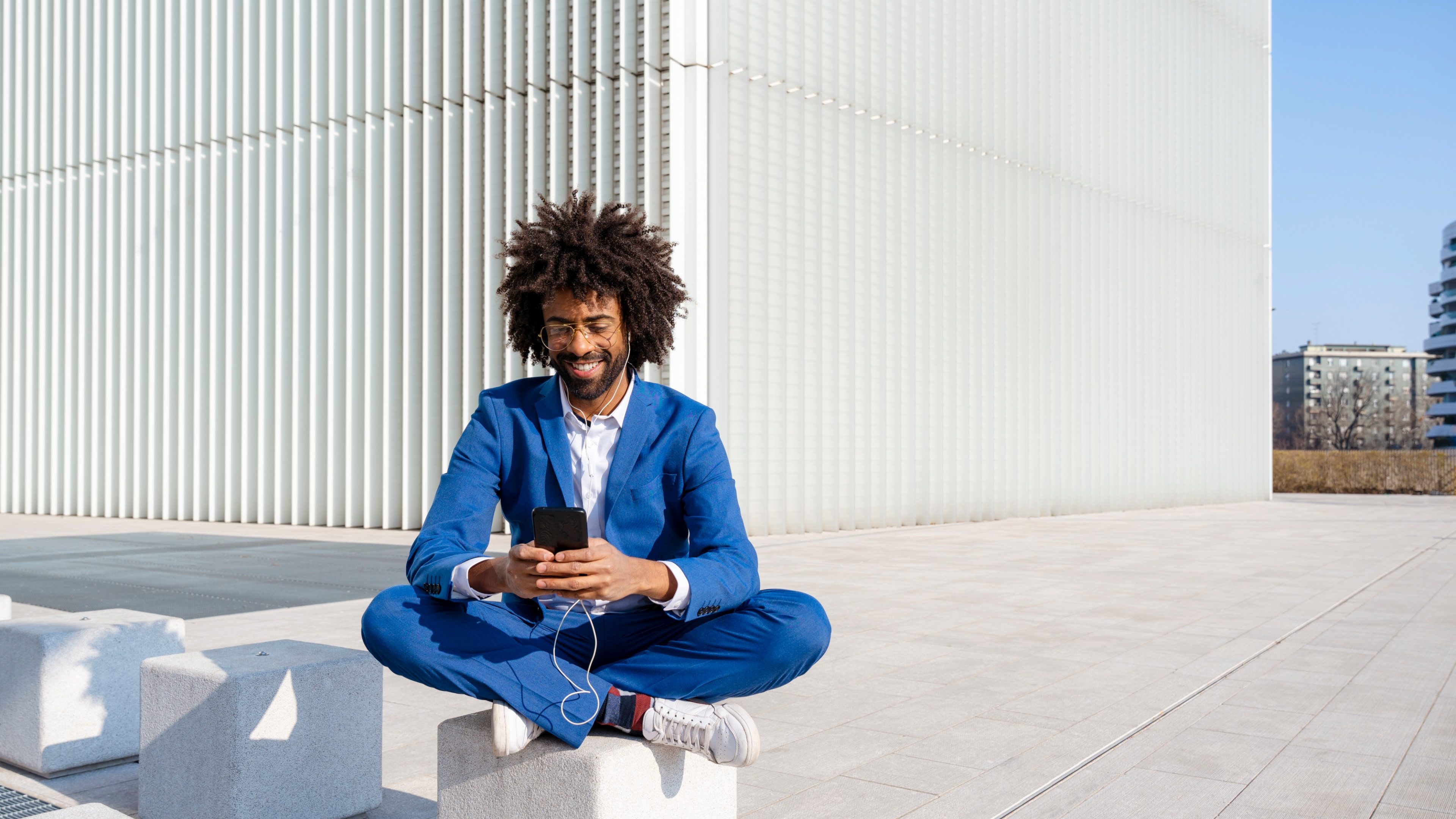 Smiling businessman listening music and using smart phone sitting on concrete block