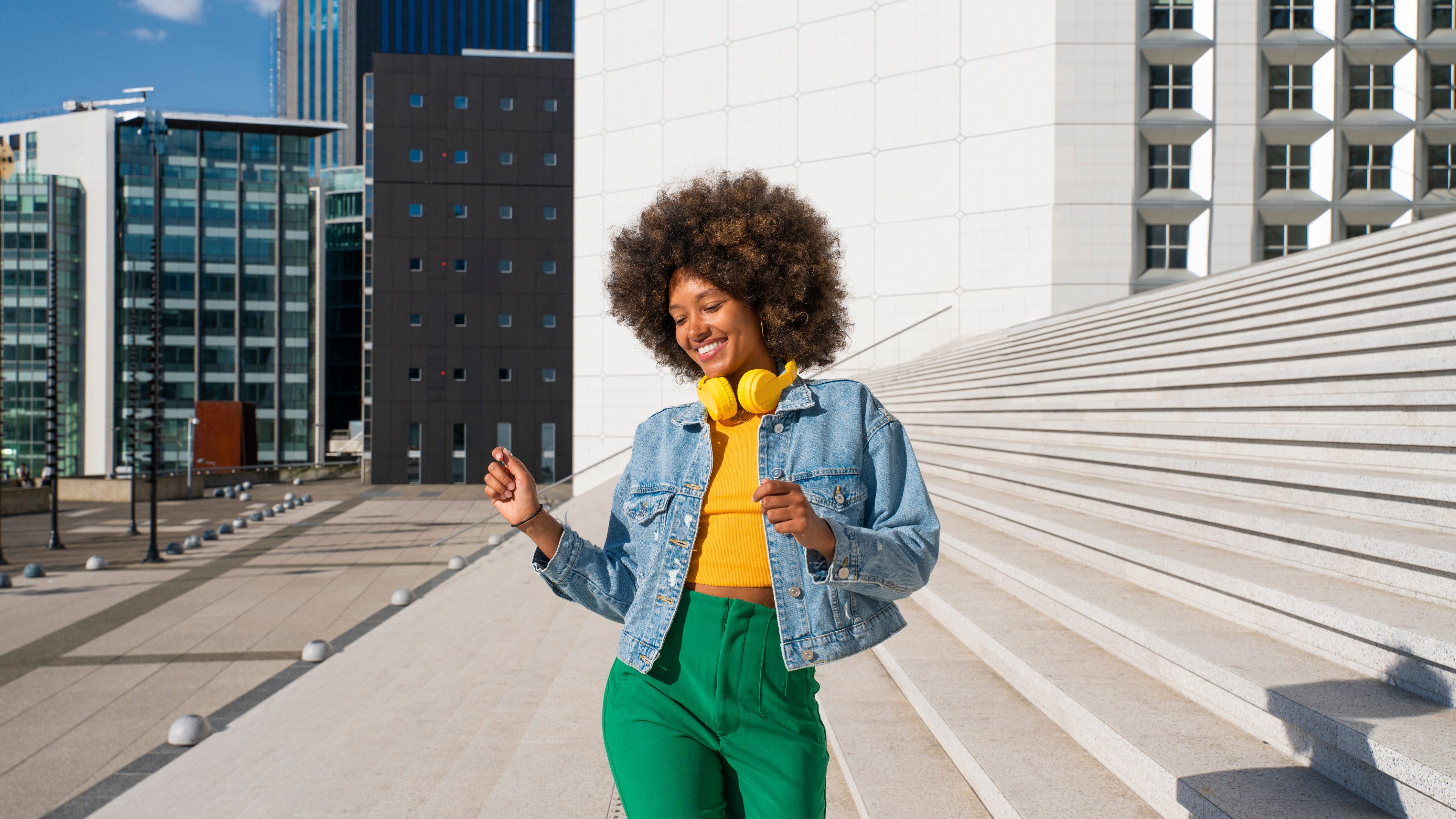 Happy Afro woman wearing wireless headphones enjoying dance by steps on sunny day