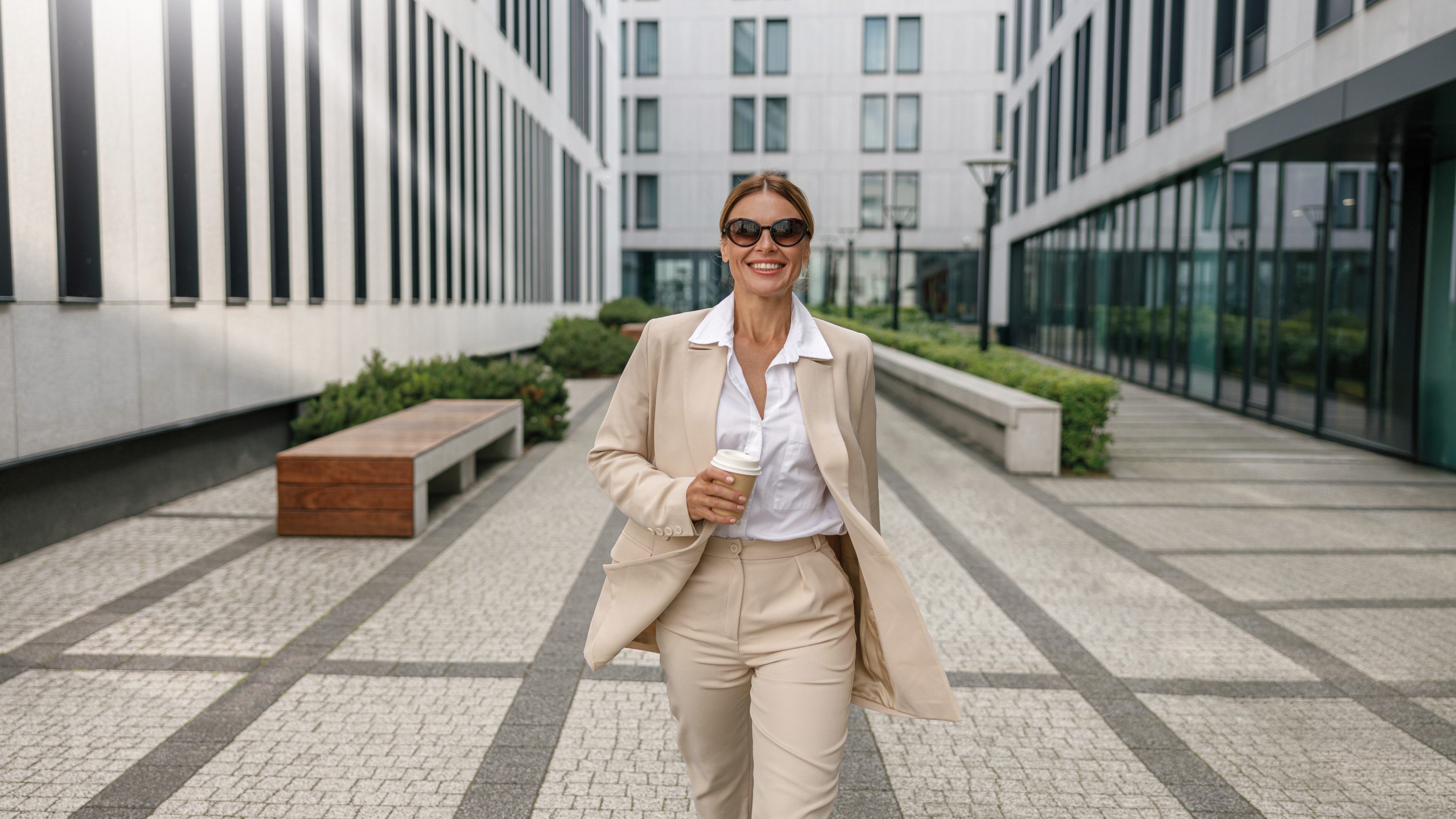 Portrait d'une femme d'affaires souriante marchant d'un pas dynamique dans un quartier de bureaux modernes, tenant un café, illustrant la vitalité et l'usage concret de l'immobilier géré par la SCPI Pierre Capitale.